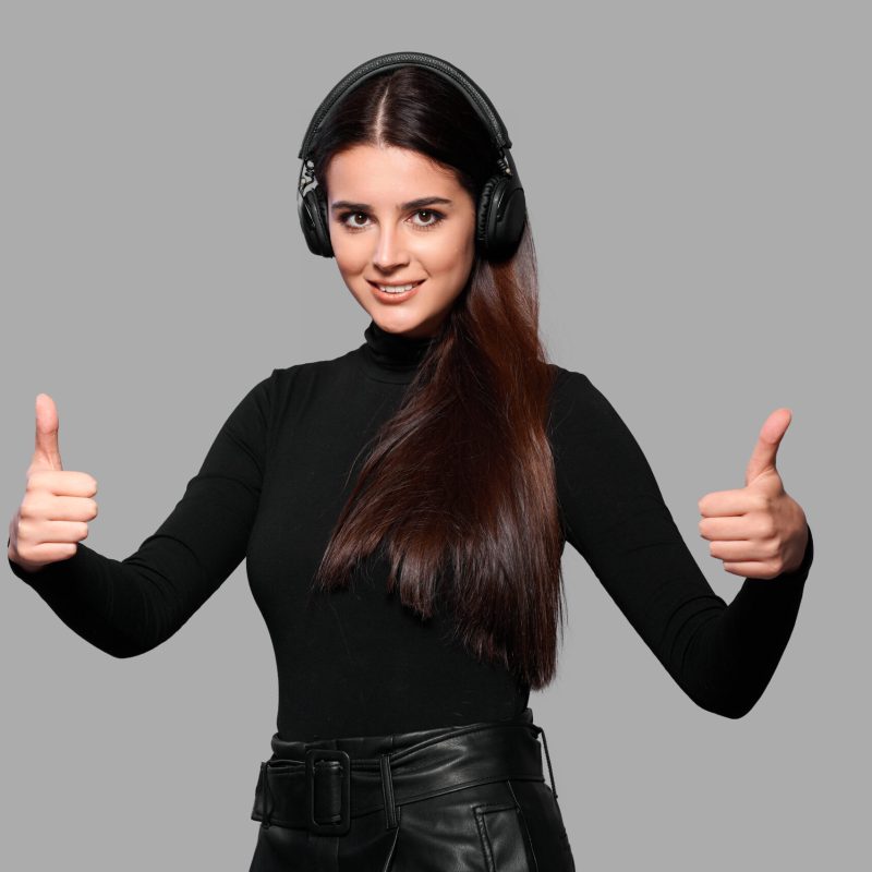 Smiling young woman in wireless headphones shows thumb up gesture, female listen to music, isolated background